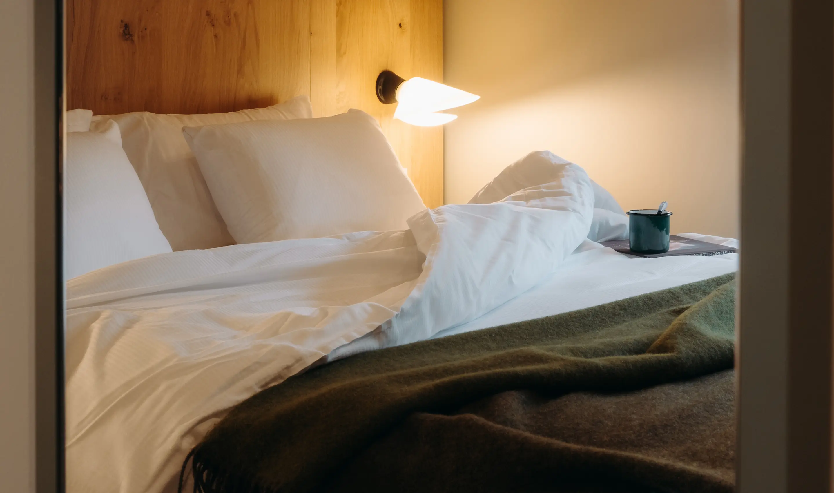 Chambre d'hôtel vue depuis l'encadrement de la porte, avec un lit double au linge blanc froissé et un plaid en laine verte. Une liseuse allumée fixée sur la tête de lit en bois éclaire chaleureusement la pièce. Sur la table de nuit, une tasse émaillée verte et un livre. Atmosphère cosy et intimiste.