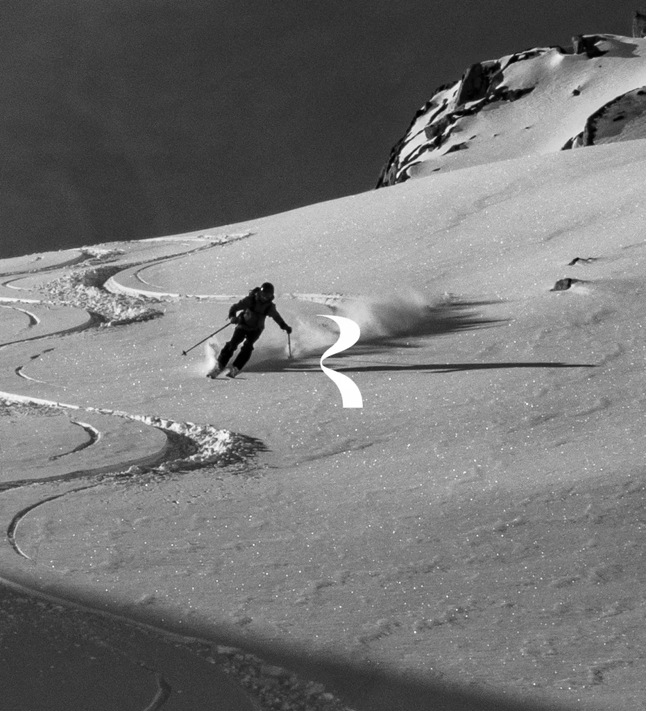 Skieur en hors-piste descendant une pente enneigée et raide en photo noir et blanc. Il trace des courbes sinueuses dans la neige poudreuse, laissant des traces visibles derrière lui. Un logo ou pictogramme blanc en forme de serpentin est superposé sur l'image. Rochers et sommet montagneux en arrière-plan.