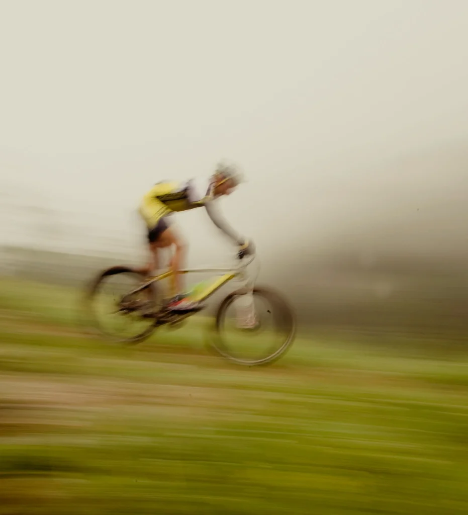 Cycliste en VTT lancé à grande vitesse sur un sentier herbeux, photographié avec un effet de filé qui accentue la sensation de mouvement. Le rider porte un maillot jaune et un casque. Arrière-plan flou de ciel gris et de prairie verte.