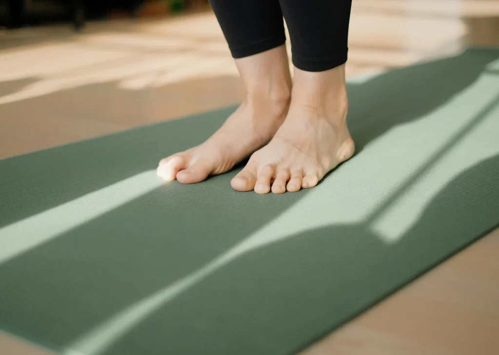 Gros plan sur les pieds nus d'une personne en legging noir debout sur un tapis de yoga vert. Des jeux d'ombres et de lumière naturelle traversent le tapis. Contexte de pratique du yoga ou du fitness en intérieur.