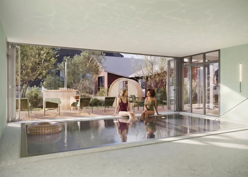 Deux femmes en maillot de bain assises au bord d'une piscine intérieure ouverte sur l'extérieur via de grandes baies vitrées coulissantes. À l'extérieur, une terrasse en bois avec des transats verts, un sauna tonneau et des arbres. Une bouée ronde flotte dans la piscine. Ambiance spa haut de gamme dans un cadre montagnard.