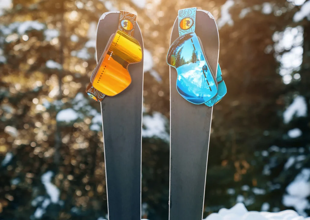 Deux paires de lunettes de ski aux verres miroir, l'une orange et l'autre bleue, posées sur les spatules de deux skis plantés dans la neige. Arrière-plan flou de sapins enneigés avec une lumière dorée de soleil couchant.