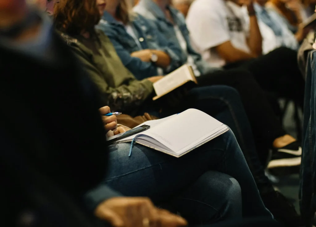 Rangée de participants assis lors d'une conférence ou d'un événement. Au premier plan, une personne tient un stylo et un carnet ouvert posé sur ses genoux, avec un smartphone. Les autres participants sont flous en arrière-plan, certains tenant également des documents ou des livres.